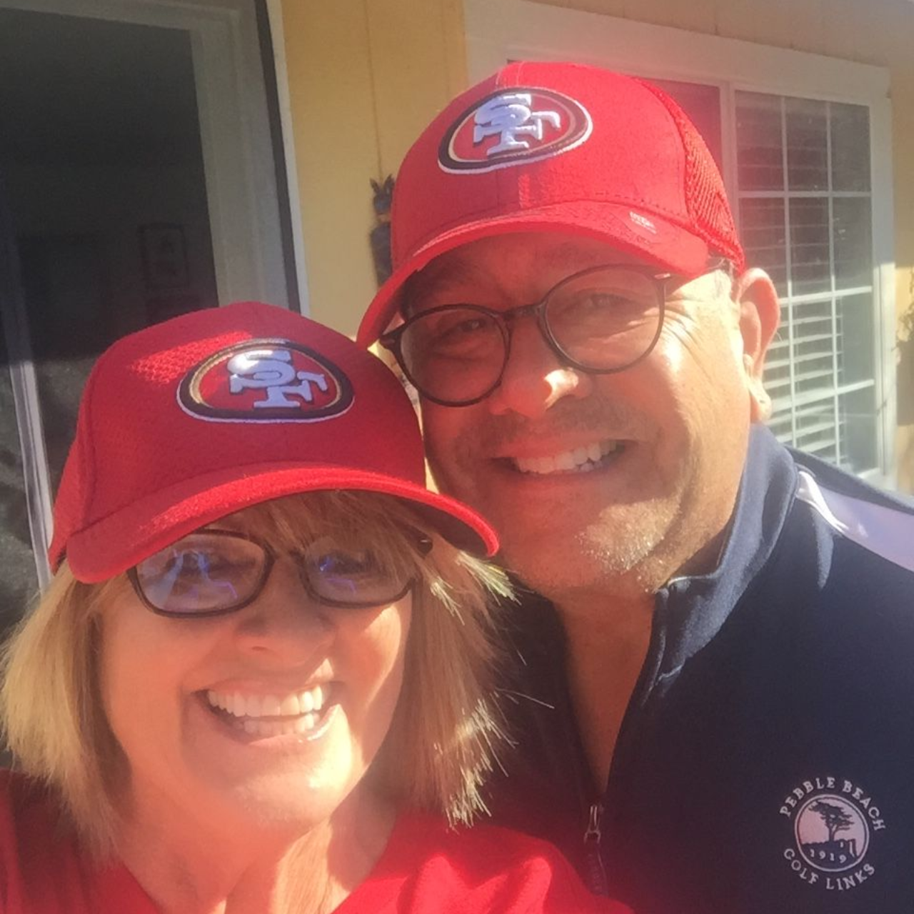 A smiling man and woman pose together wearing red San Francisco 49ers baseball caps.