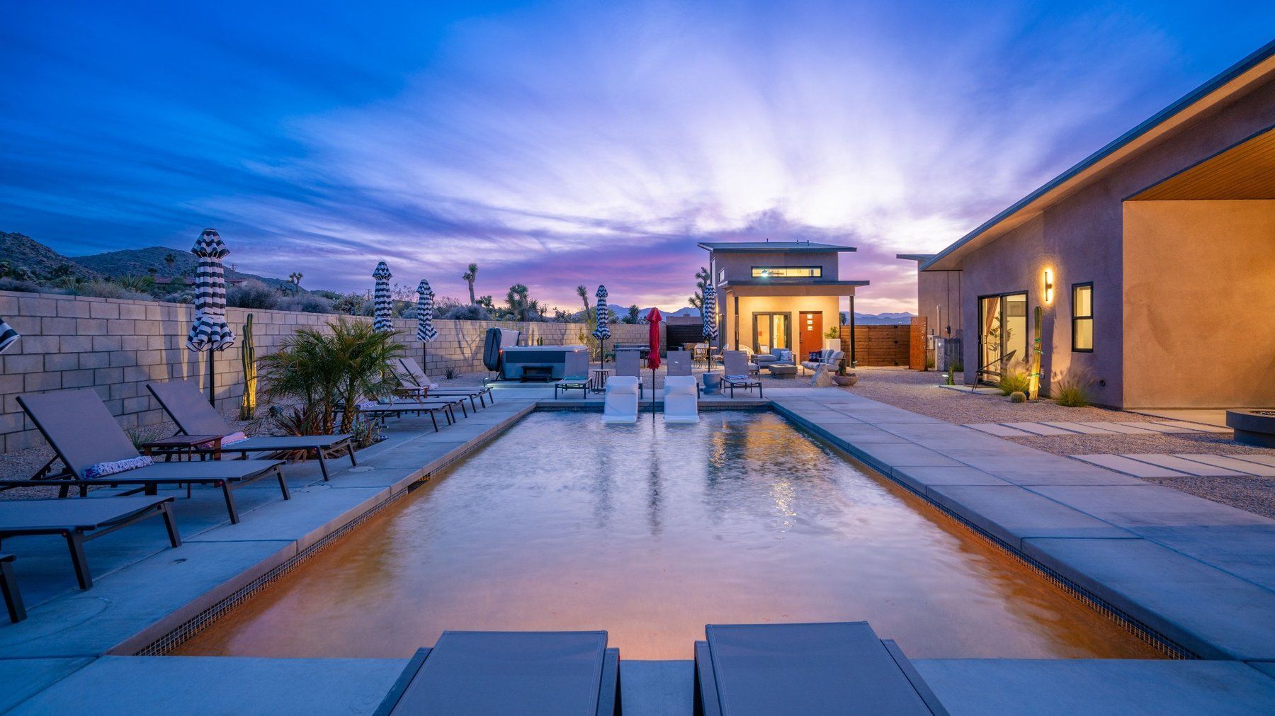 Modern rooftop pool at dusk with lounge chairs and city skyline beyond water.