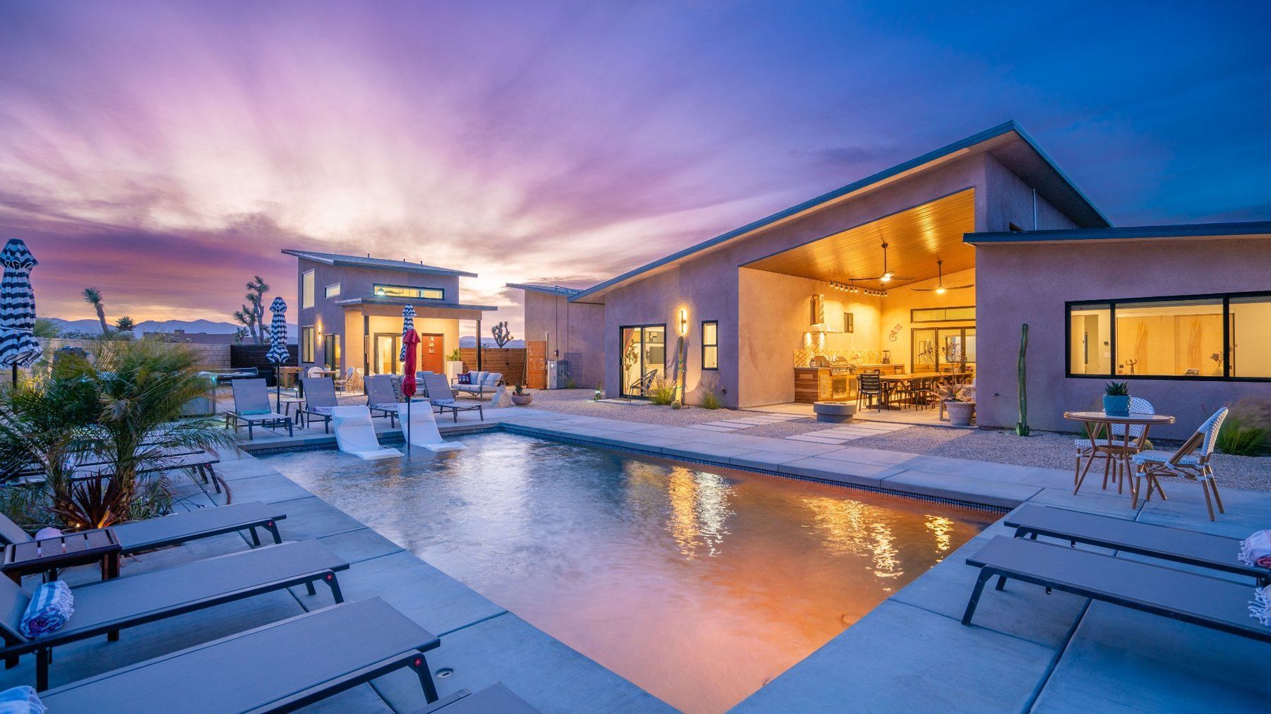 Modern outdoor patio with a lit swimming pool at sunset, surrounded by lounge chairs and a contemporary house