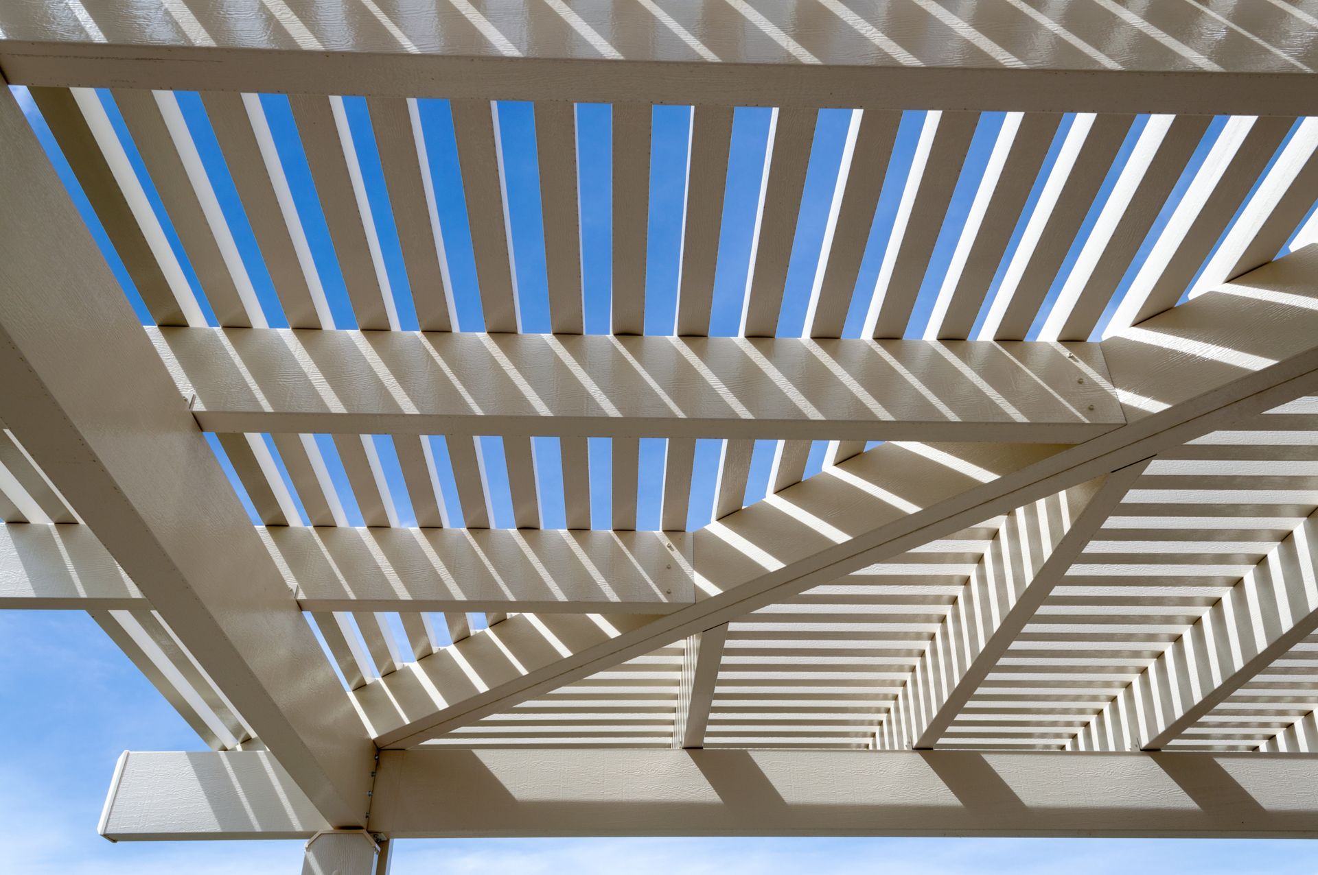 Sunlight shining through the slatted beams of a pergola against a clear blue sky.