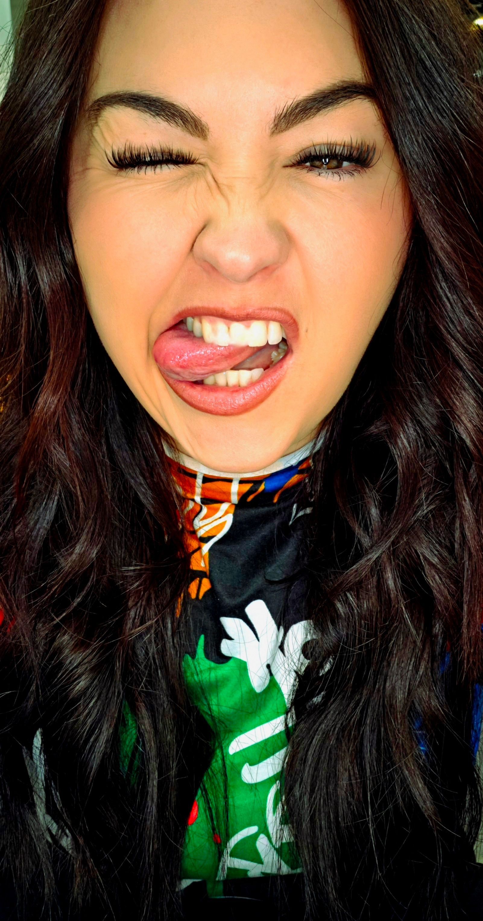 Close-up of a person making a playful face with tongue out, wearing a green shirt and black hair.