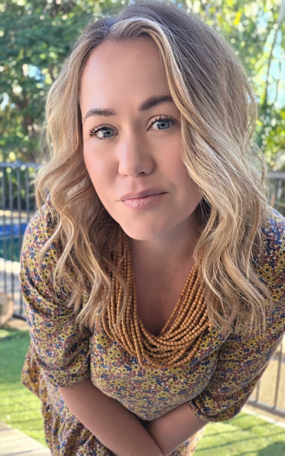 Close-up portrait of a smiling woman with wavy blonde hair in a patterned top outdoors, with greenery behind her