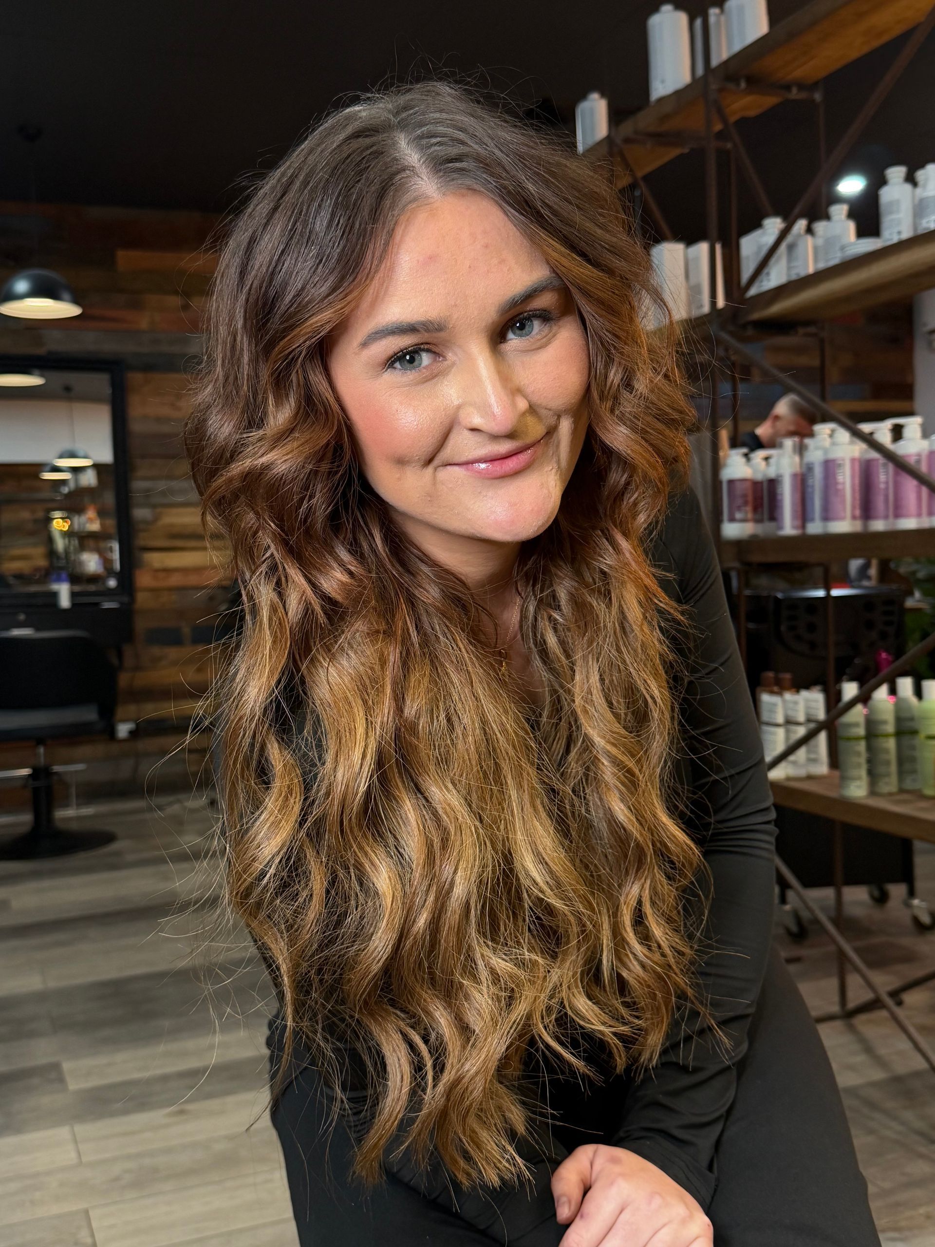 Woman with long wavy brown hair smiling in a modern café interior.