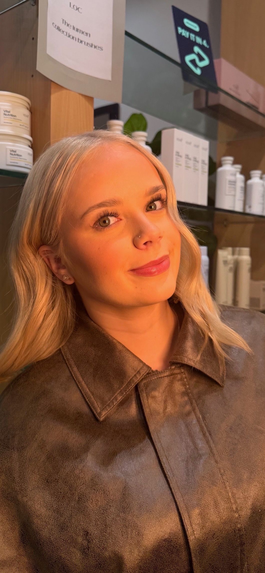 Woman with blonde hair in a brown sweater, smiling indoors beside shelves of skincare products