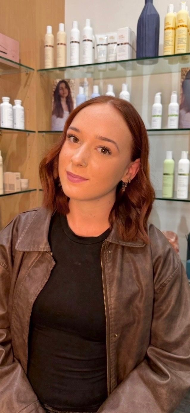 Woman with dark hair in a black top and brown jacket, standing in a salon with hair products on shelves