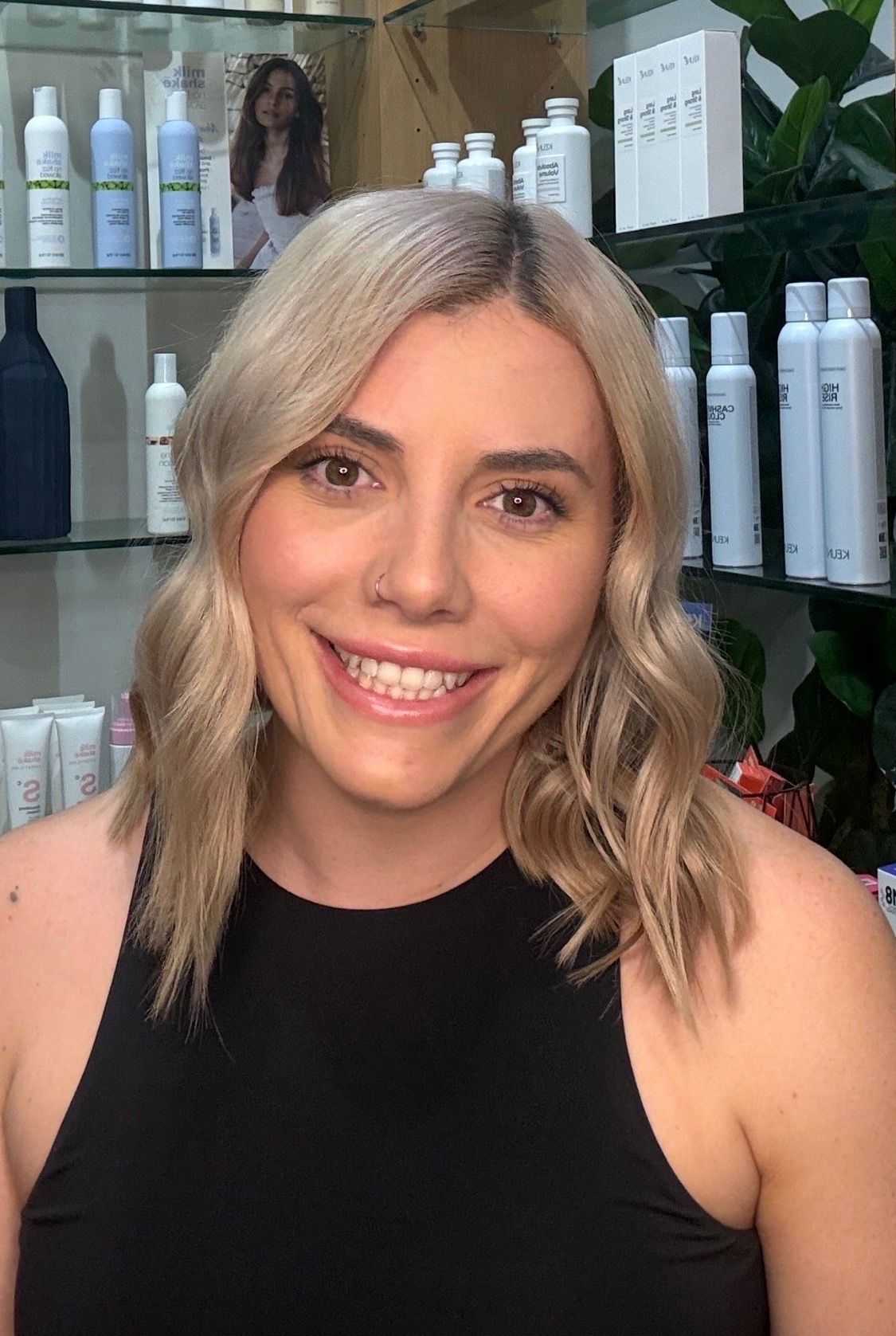 Smiling woman with blonde hair in a black sleeveless top, standing in front of salon product shelves.
