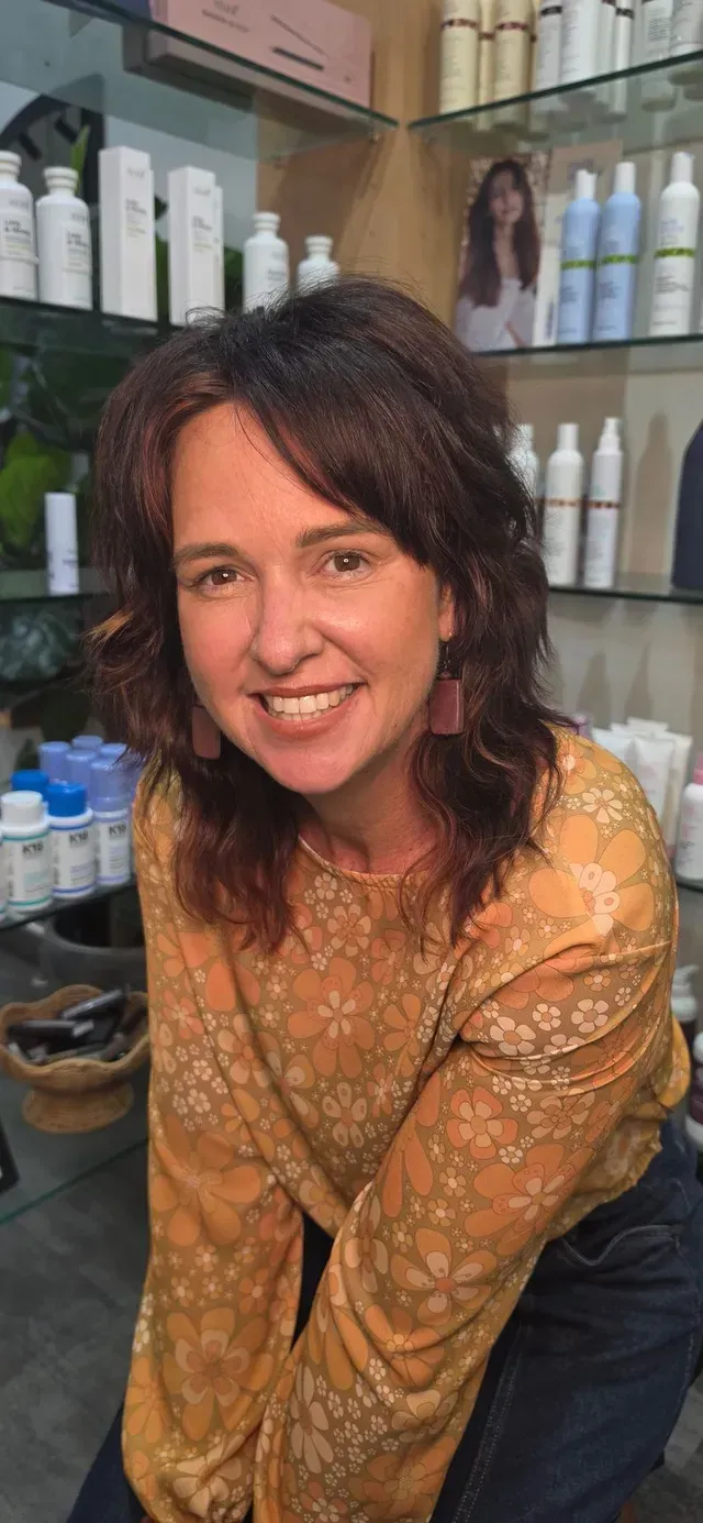Woman smiling in a salon, leaning forward in a patterned yellow top with hair products on shelves behind her.