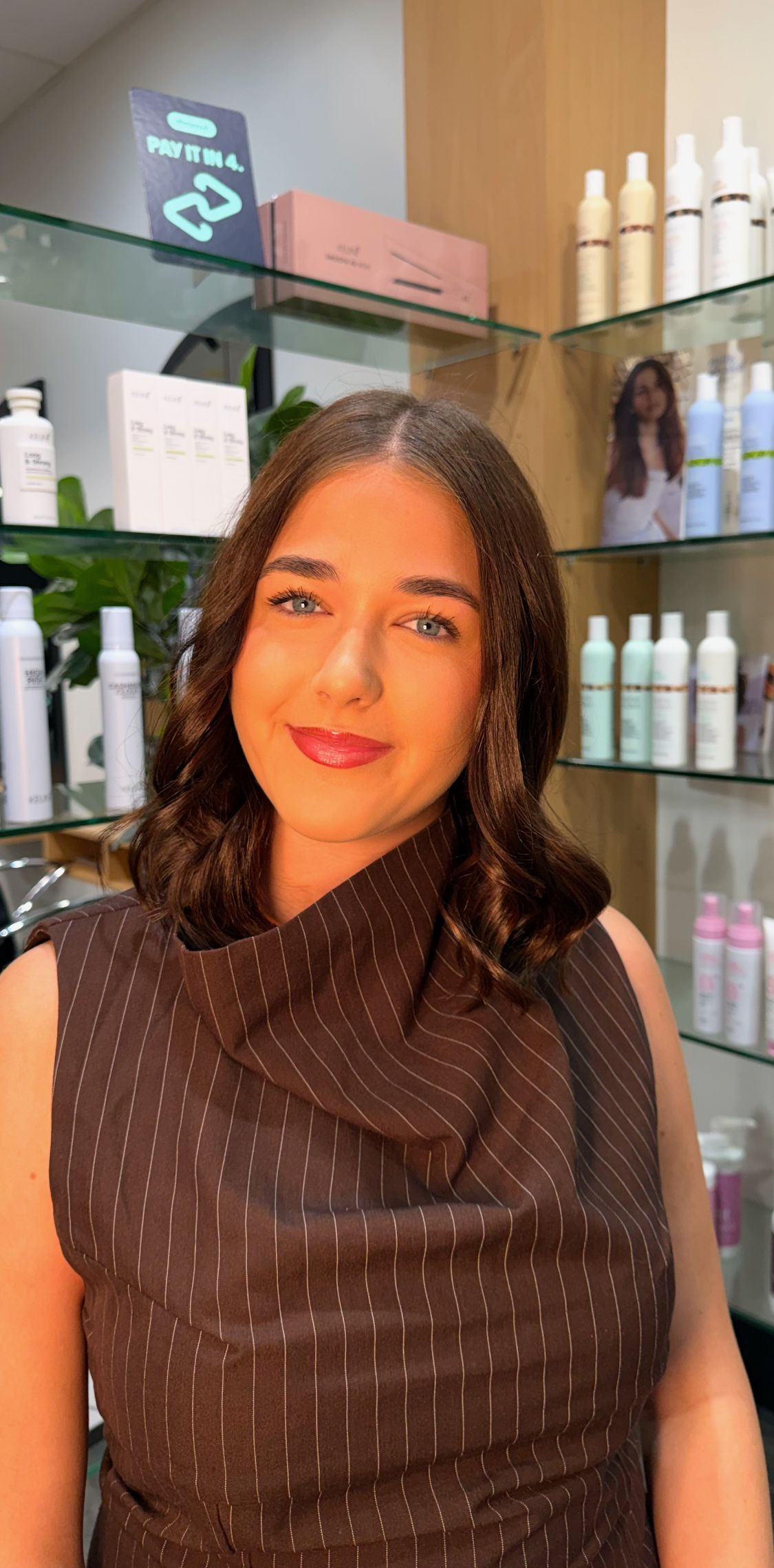 Woman smiling in a salon, with shelves of hair and skincare products behind her.