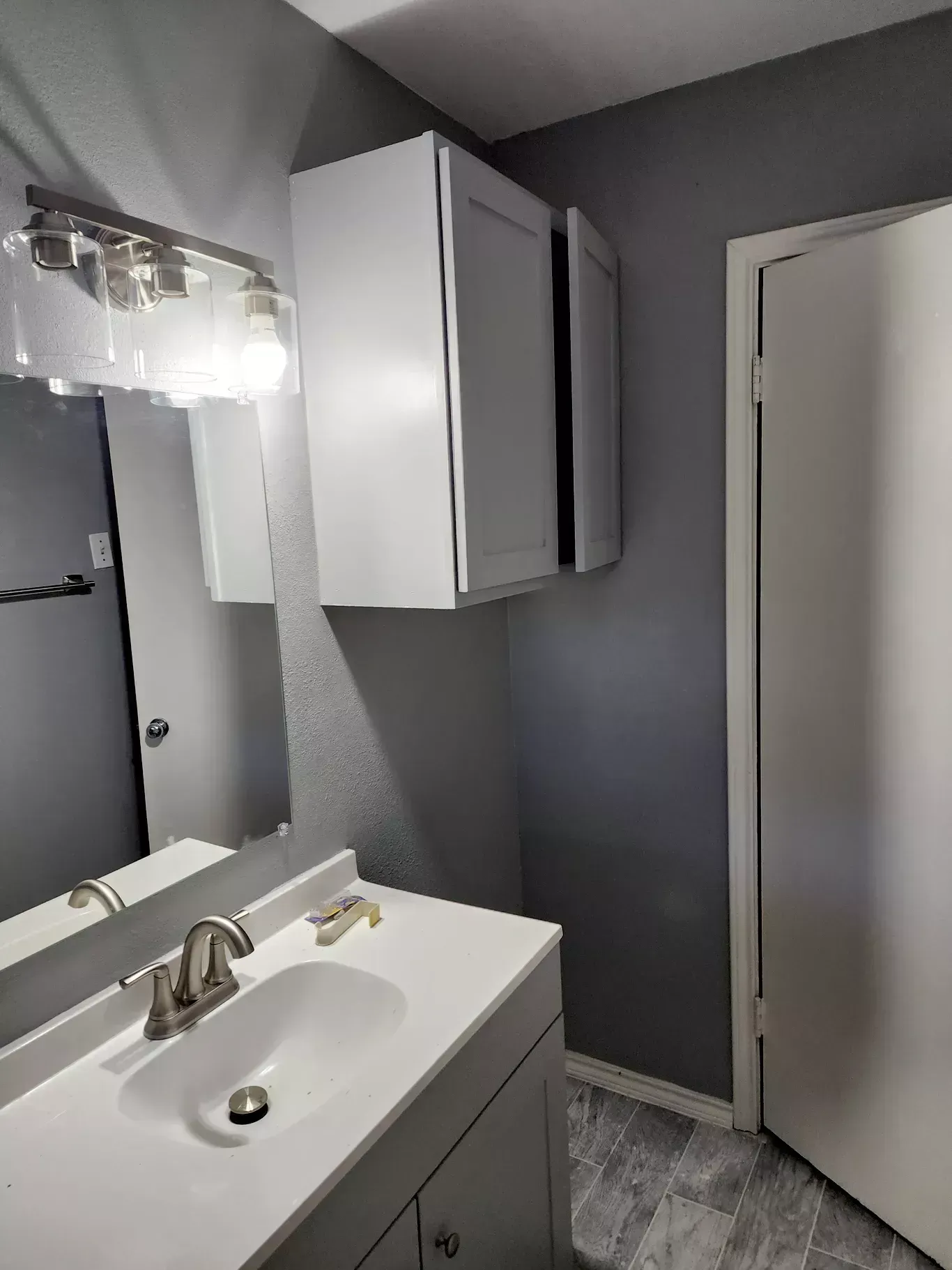 Small bathroom vanity with mirror, gray walls, cabinet, and open door.