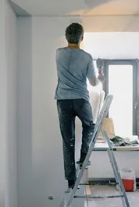 Person standing on a ladder painting a white wall near a window in a room.