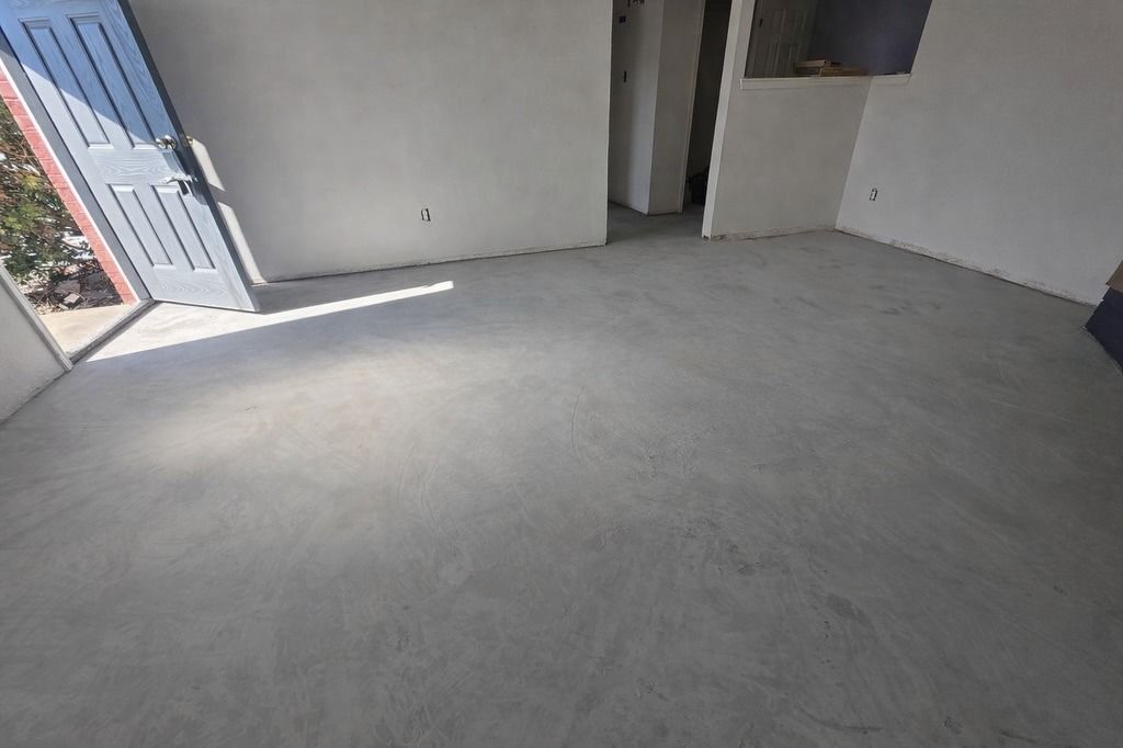 Empty unfinished room with gray floor, white walls, and sunlight from an open doorway.