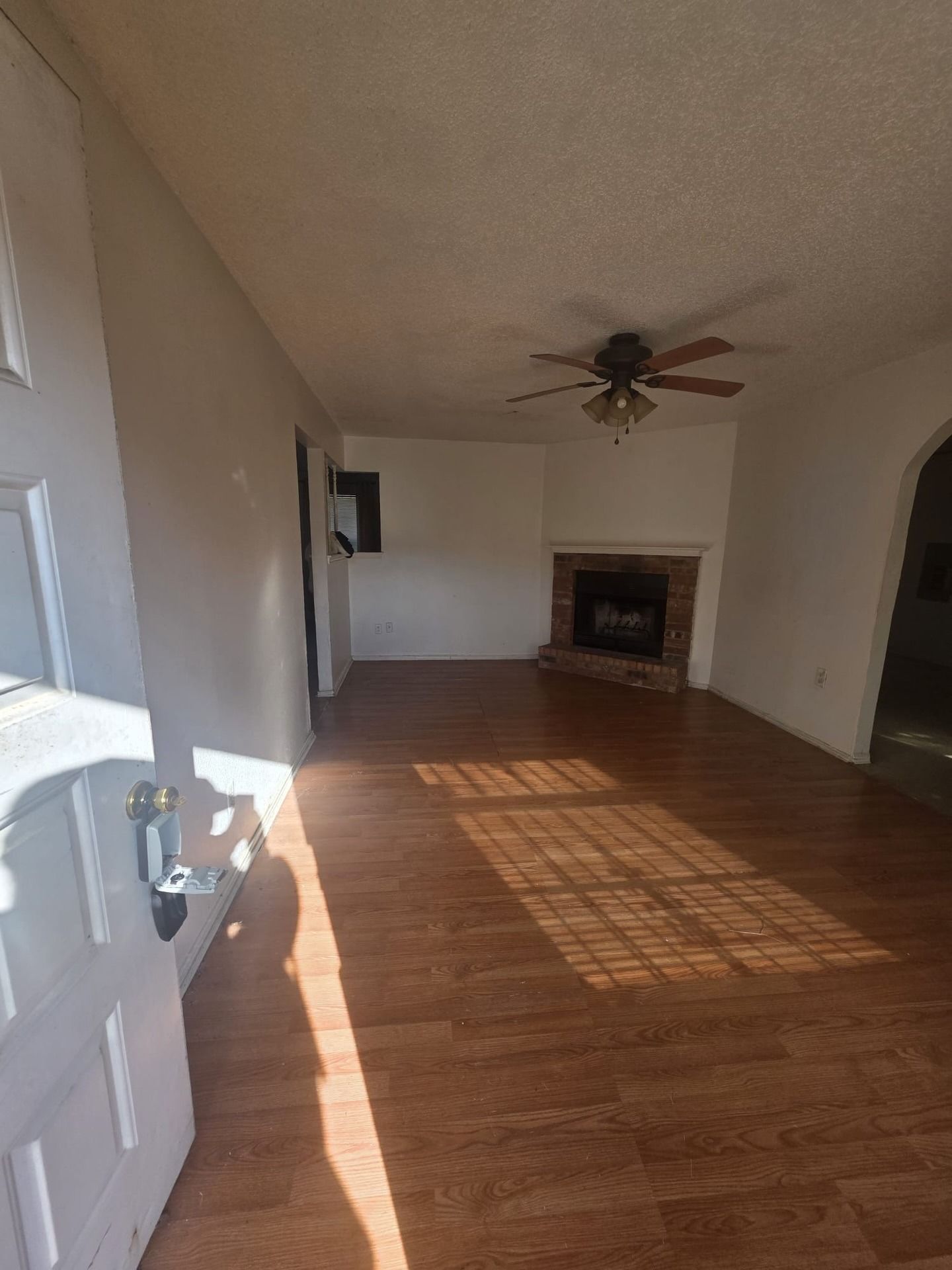Empty living room with hardwood floors, ceiling fan, and fireplace, lit by sunlight through a window.