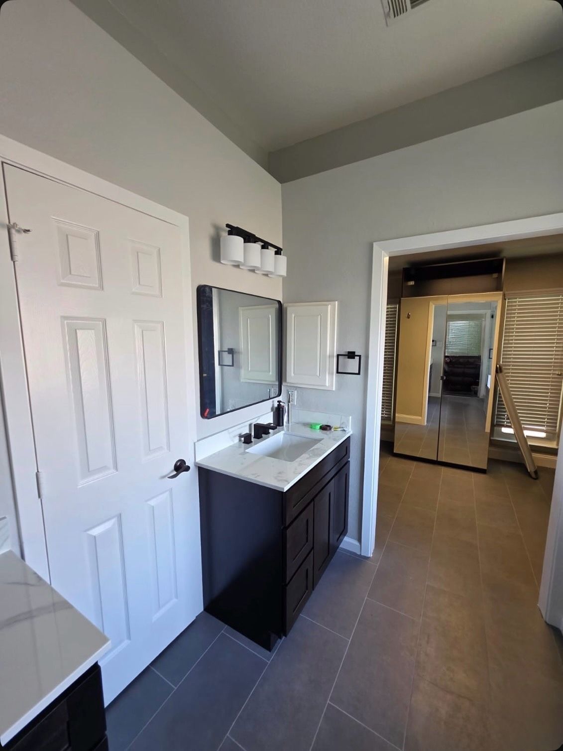 Modern bathroom with dark vanity, white door, wall-mounted mirror, and gray tile floor leading to adjoining space