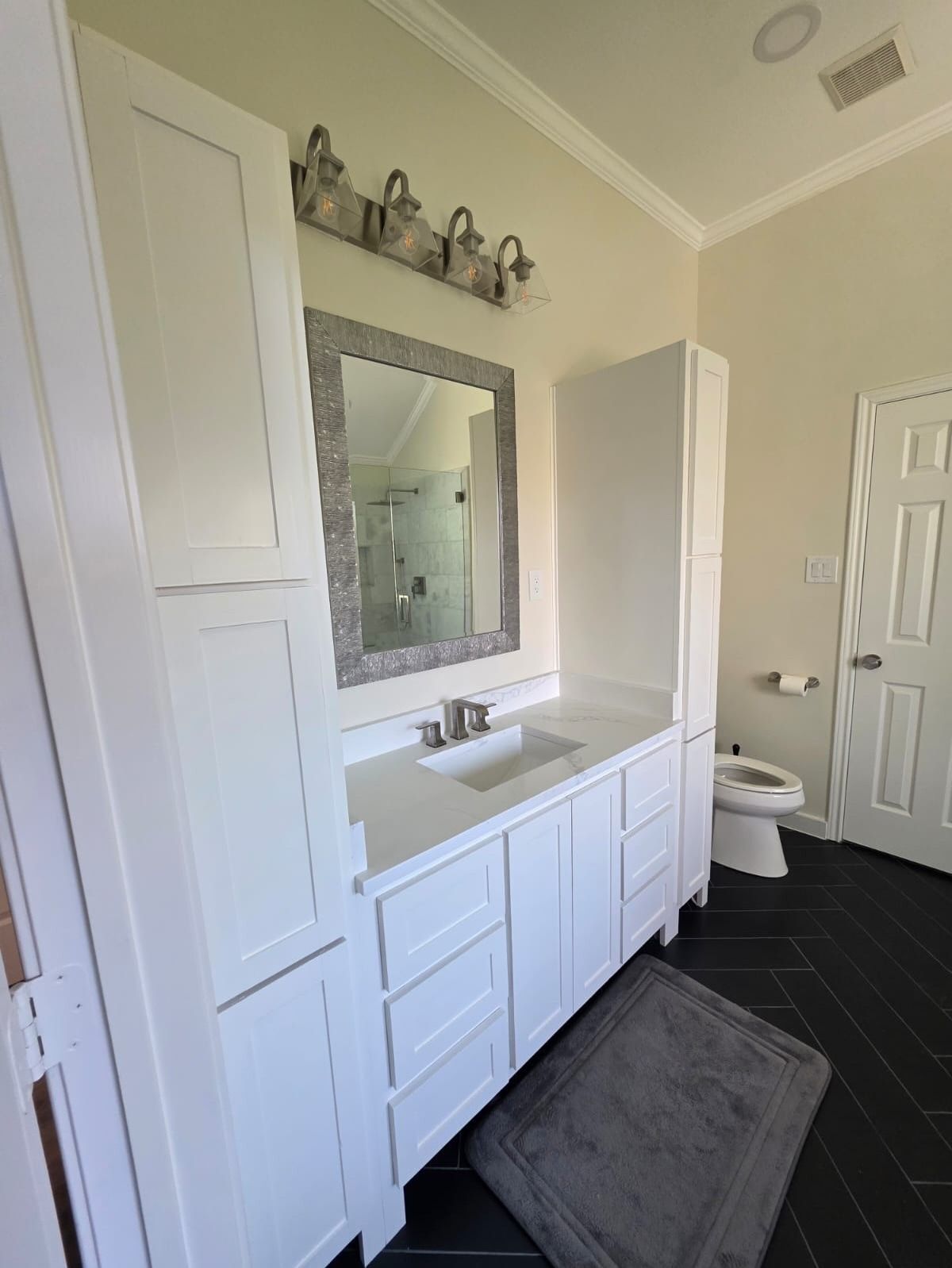White bathroom vanity with mirror, wall sconces, dark floor, and gray bath mat near a toilet