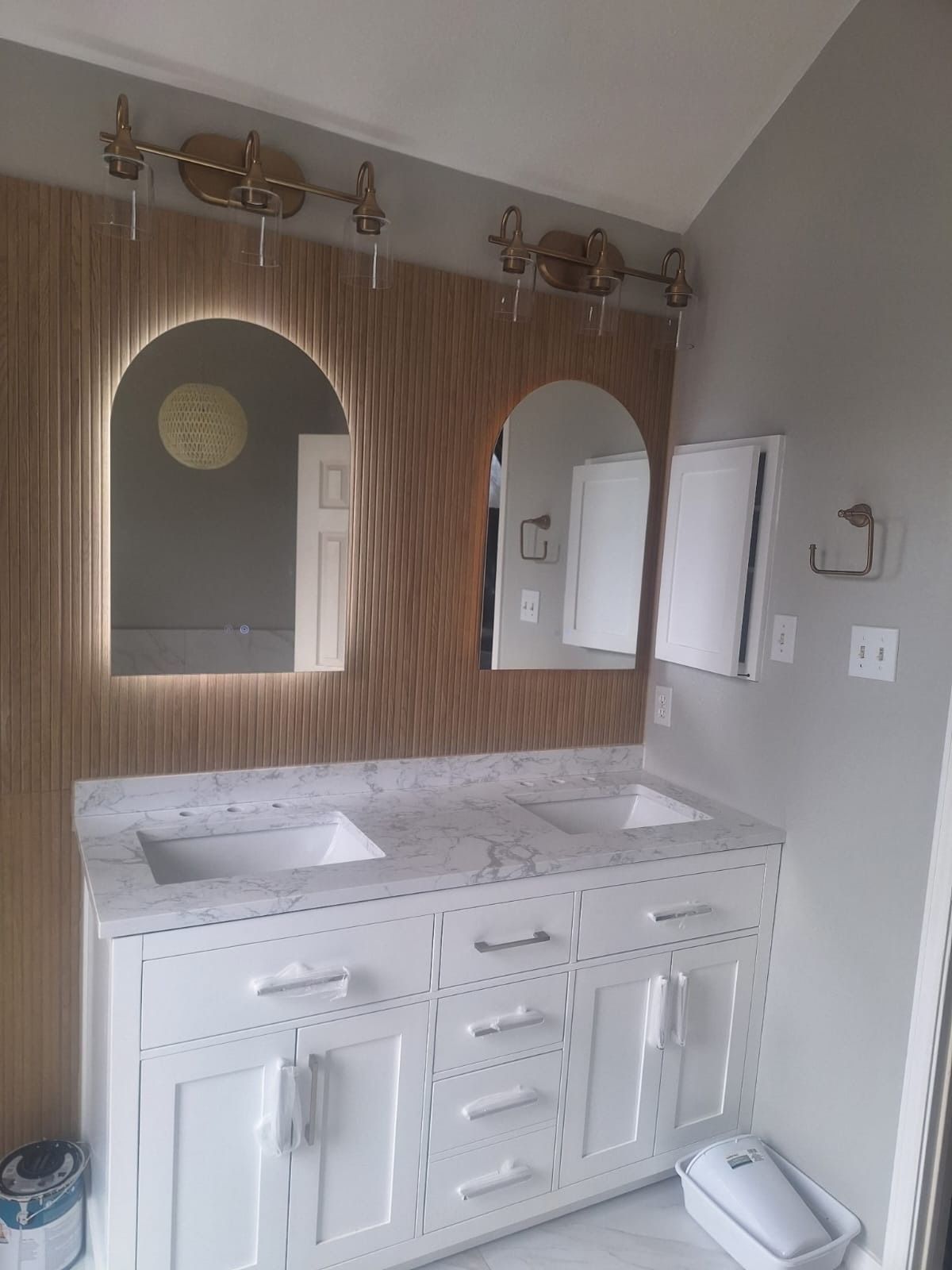 Bathroom vanity with marble countertop, white cabinets, arched mirrors, and wall sconces against a wood accent wall