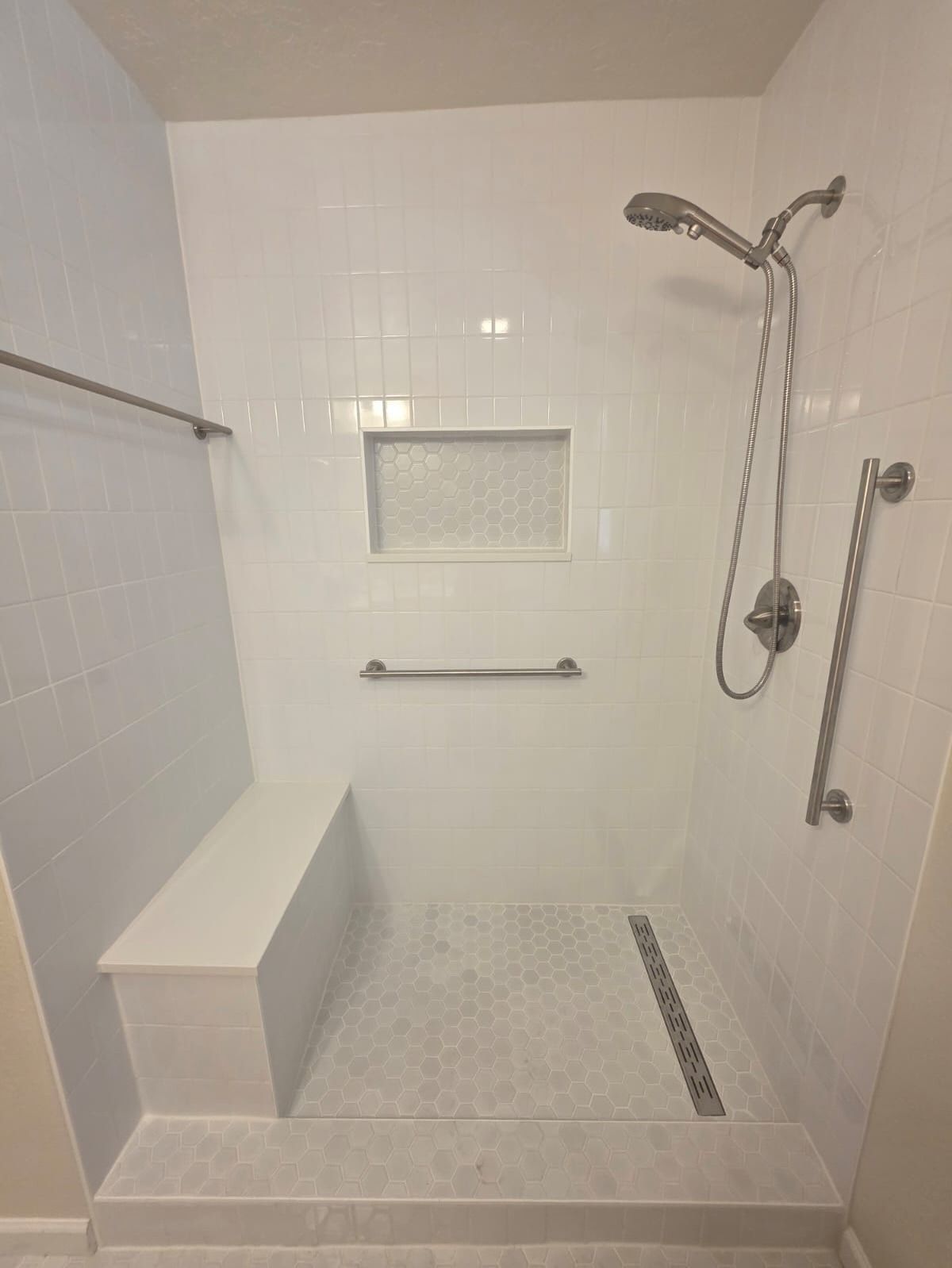 Accessible white tiled walk-in shower with built-in bench, grab bars, handheld showerhead, and niche