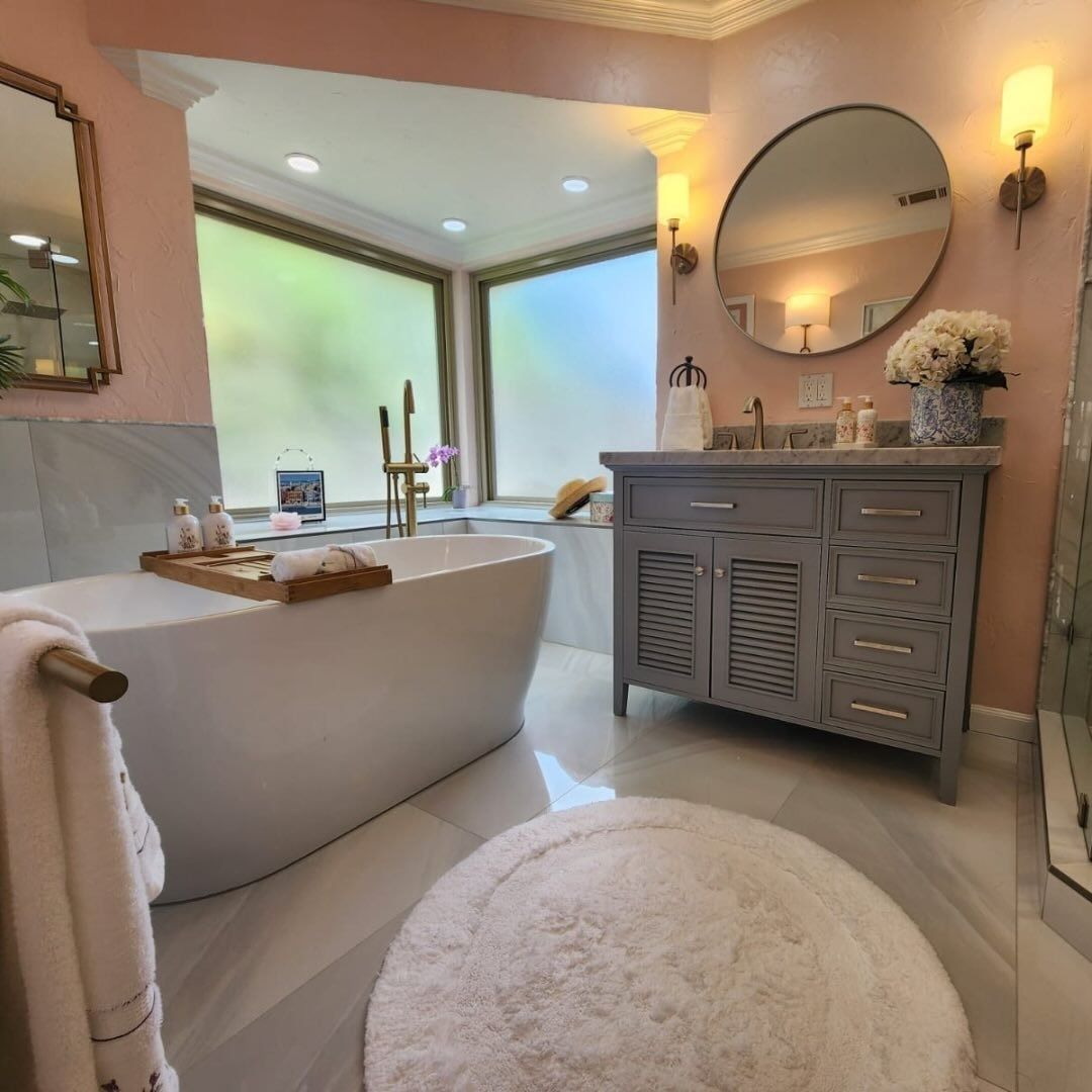 Elegant pink bathroom with a freestanding tub, vanity, round mirror, and large corner windows
