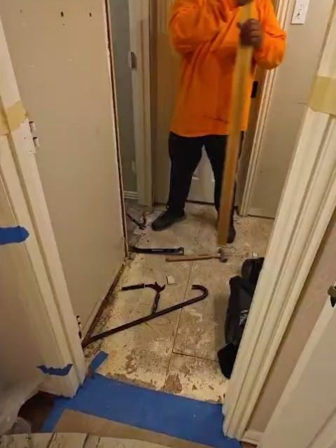 Person in orange sweatshirt standing in a damaged hallway with debris and a broken floor board