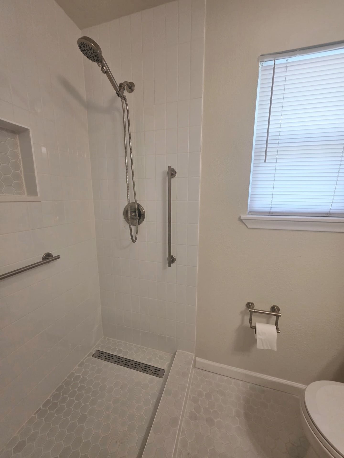 Walk-in shower with bench, handheld showerhead, grab bar, and window beside a toilet.