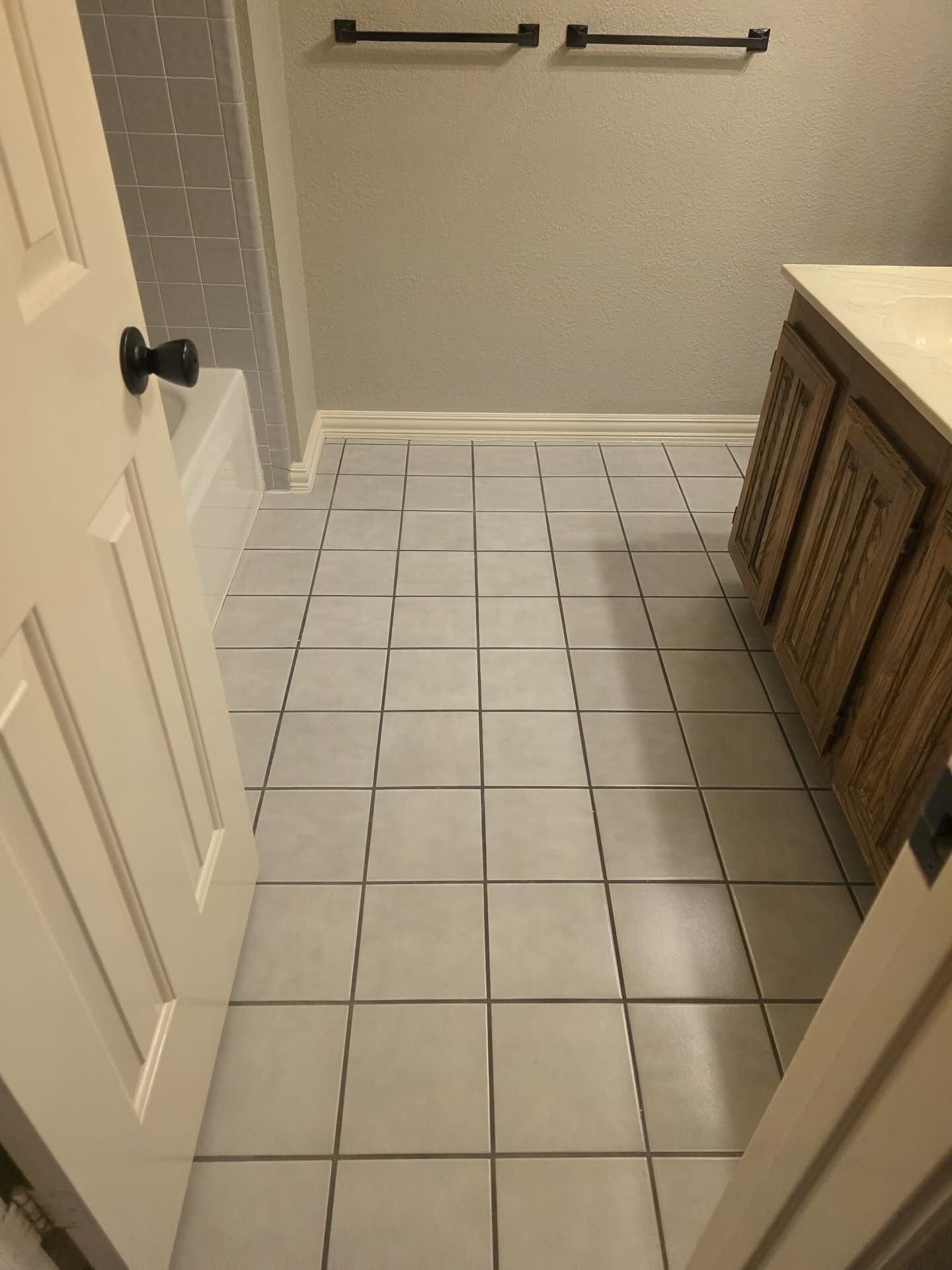 Small bathroom with white tiled floor, beige walls, a vanity, towel bar, and an open door.