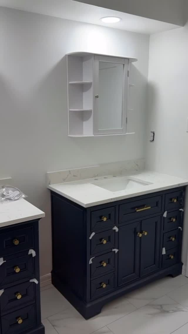 Bathroom vanity with dark blue cabinets, white countertop, and mirrored wall cabinet in a small white room