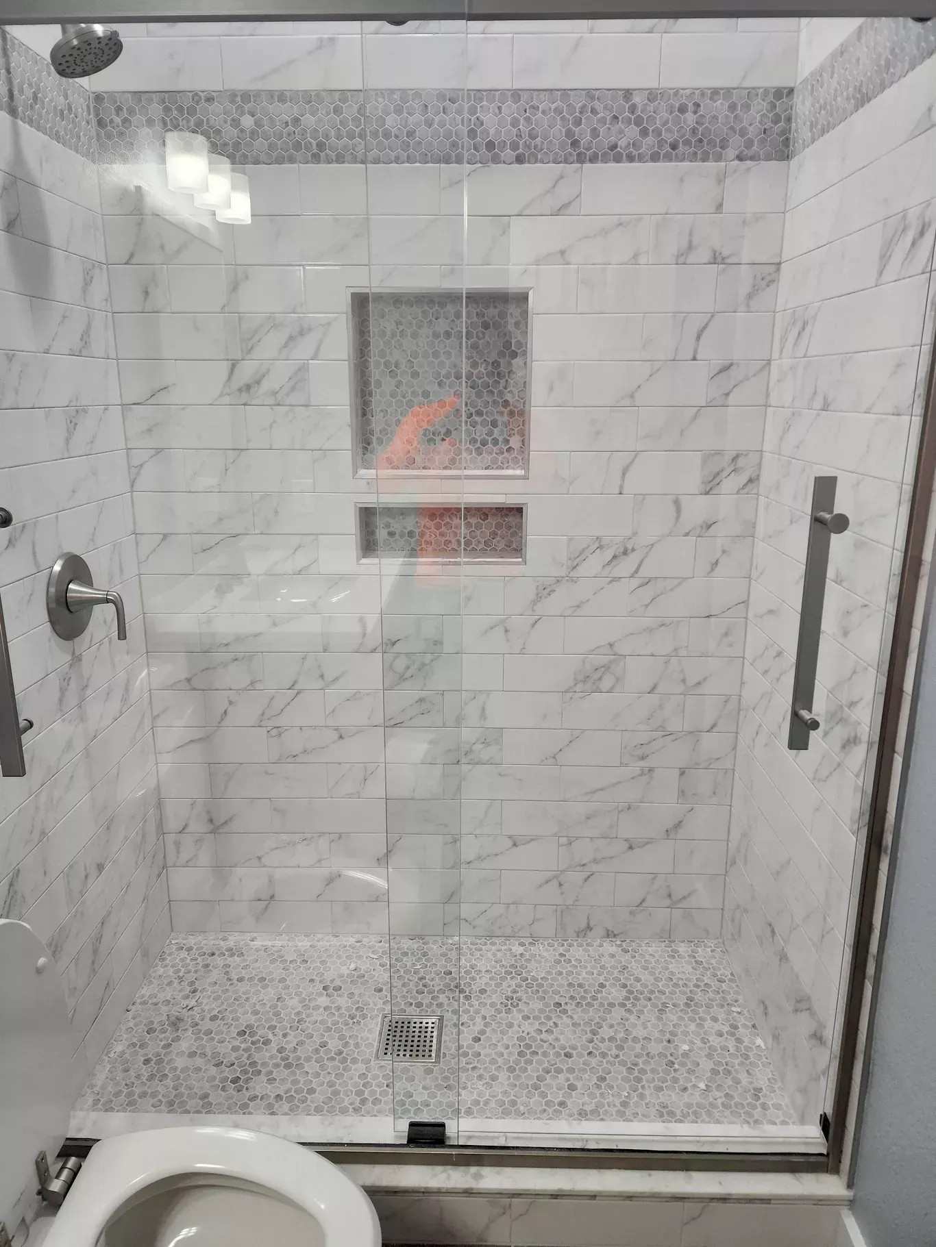 White tiled glass shower stall with built-in shelf and grab bar, viewed from outside.
