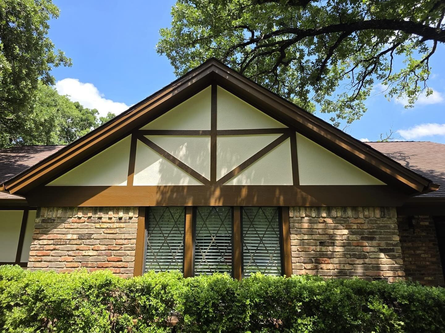 Brick house facade with a peaked roof, green shutters, and trimmed hedges under trees