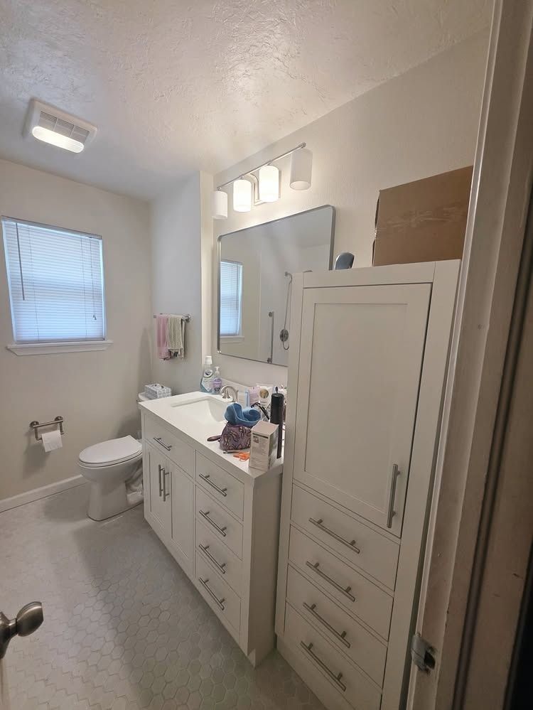 Small bathroom with white vanity, toilet, shower, window, and mirrored cabinet in a beige interior.