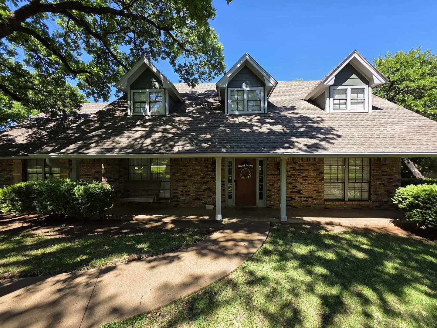 Brick house with a covered porch, dormer windows, and a sunlit front yard under trees.