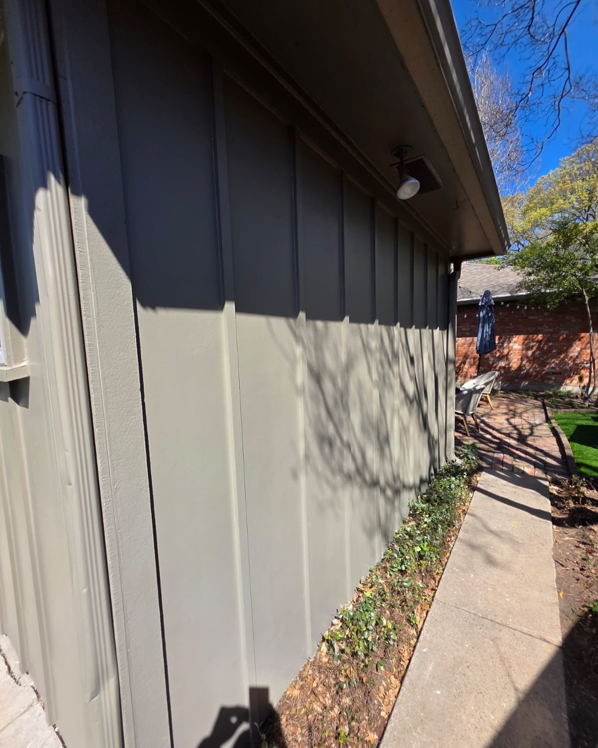 Side of a beige house beside a concrete walkway, with shadows and a brick wall in the background