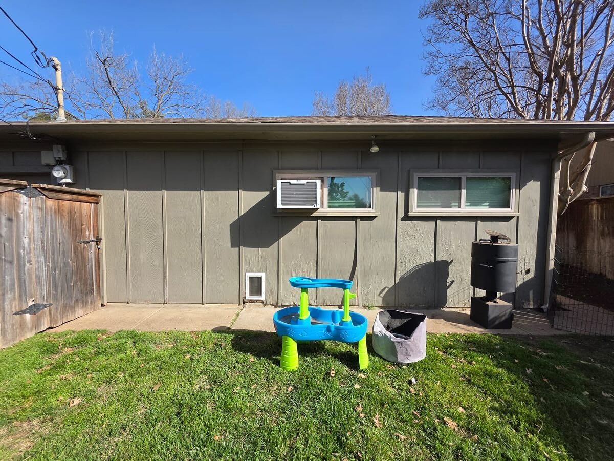 Backyard shed wall with small windows, green grass, blue child’s play table, and a black grill under a clear sky