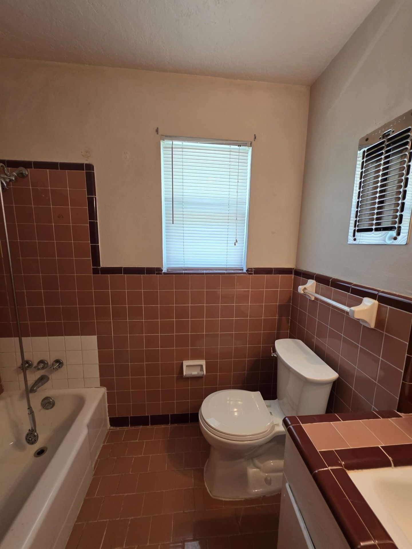 Small bathroom with brown tile, bathtub, toilet, sink, and a window with blinds.