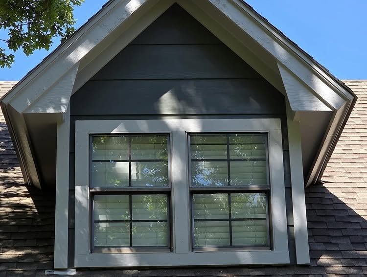 Dormer window with white trim on a gray house beneath a peaked roof, lit by sunlight