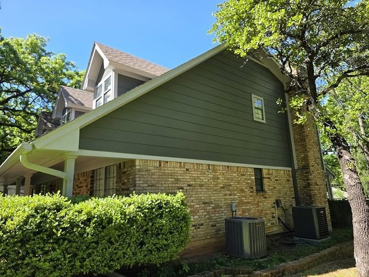 Side view of a brick house with green siding, large trees, and a sunny blue sky