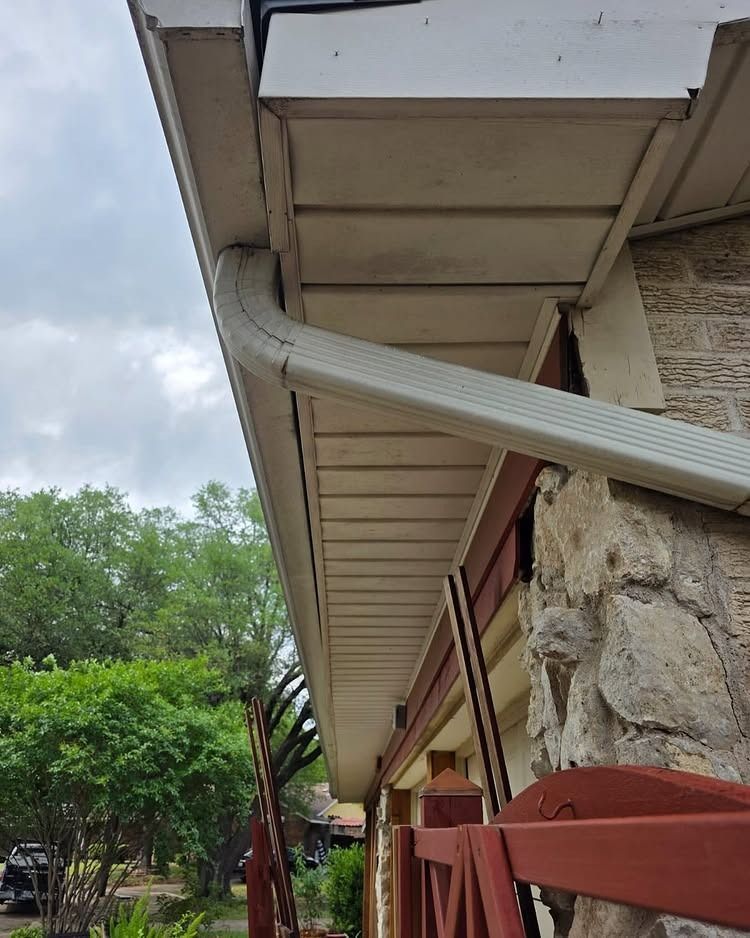House eaves with white gutter and downspout beside a stone wall, under a cloudy sky.