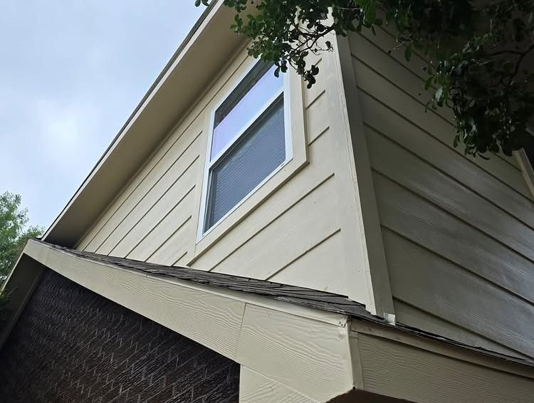 Exterior corner of a beige house with a white-trimmed window and roofline viewed from below