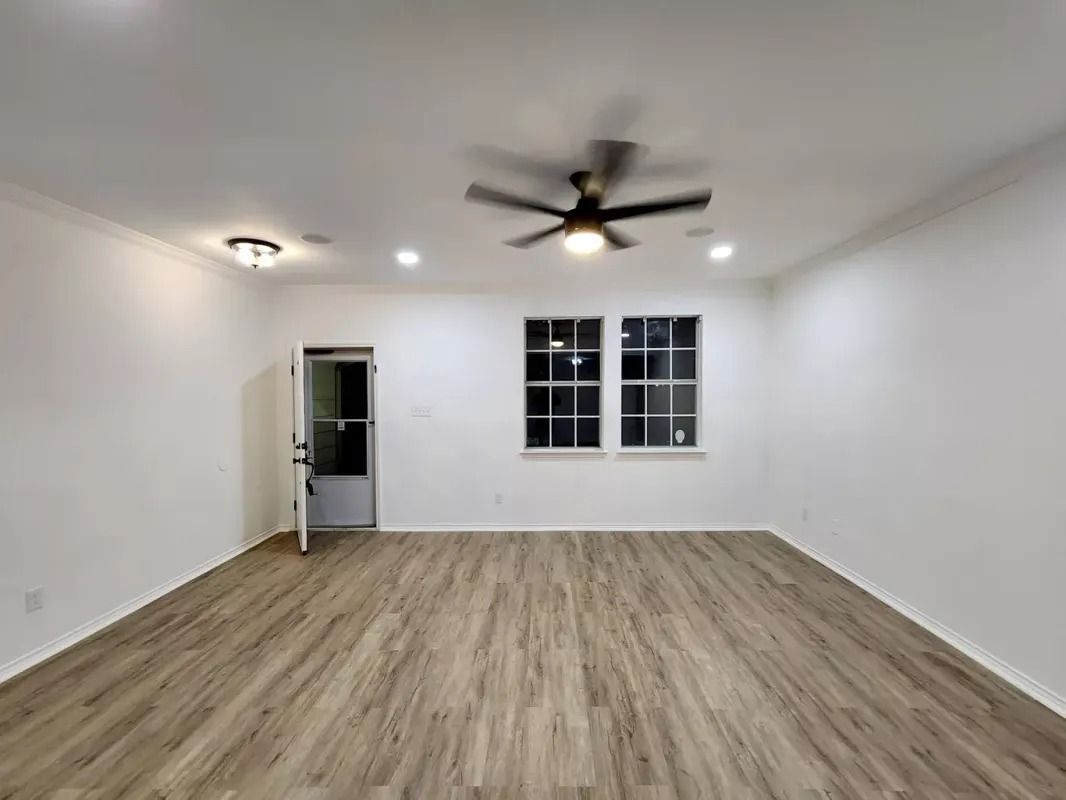Empty white room with wood-look flooring, ceiling fan, recessed lights, and windows near an open door.