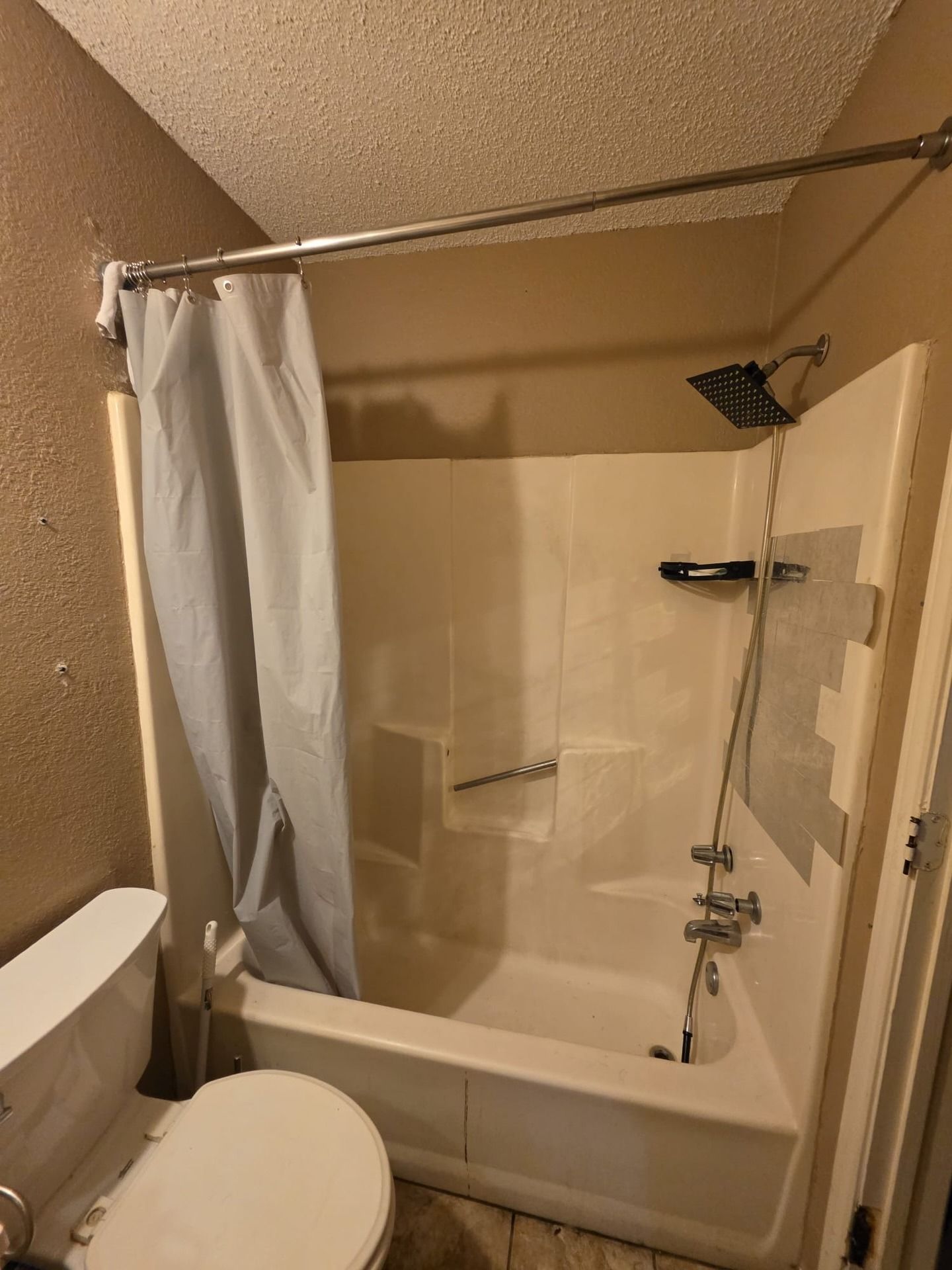 Small beige bathroom with tub-shower, white curtain, showerhead, and toilet in the foreground