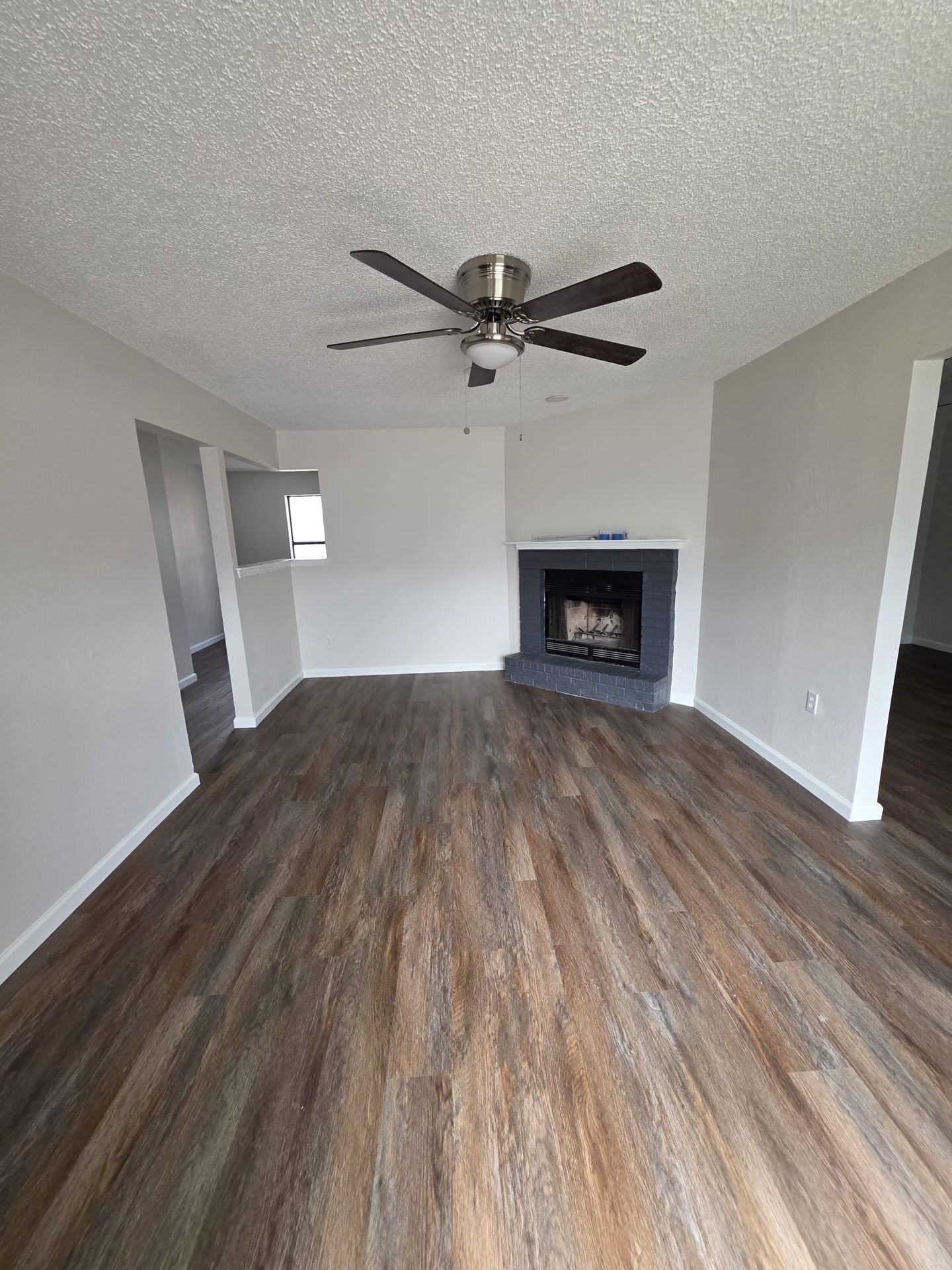 Empty living room with wood-look floors, white walls, ceiling fan, and a fireplace.