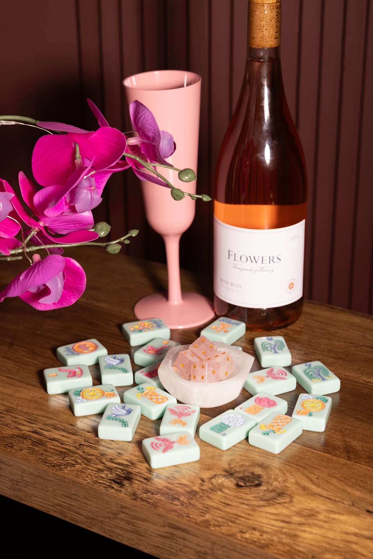 Wine bottle, pink glass, orchid, and pastel soaps arranged on a wooden table.