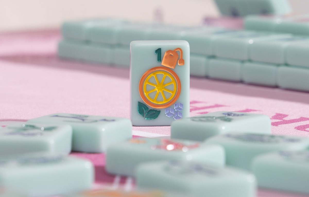 Mahjong tile with orange fruit design standing among pale blue tiles on a pink surface