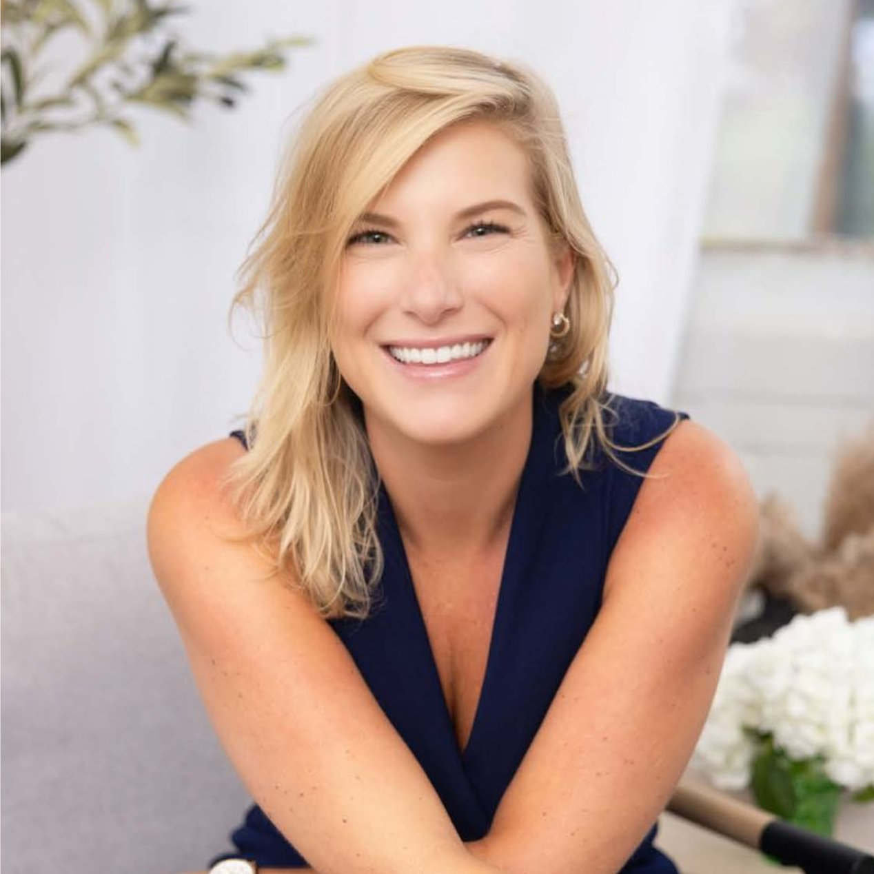 Smiling woman in a navy sleeveless top leaning forward in a bright, softly blurred room