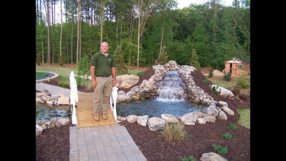 Man standing on a wooden bridge beside a rock waterfall in a landscaped garden