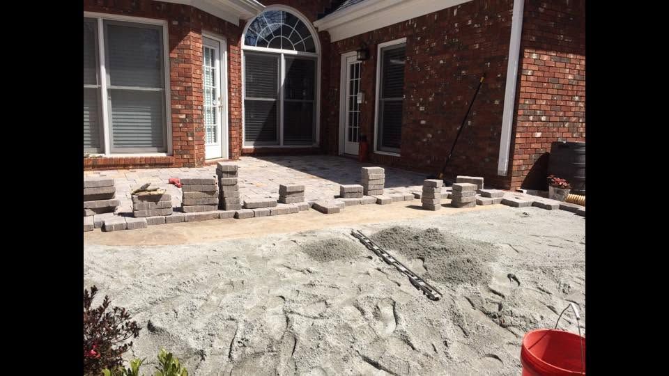 Stone patio under construction in front of a brick house with stacked pavers and tools