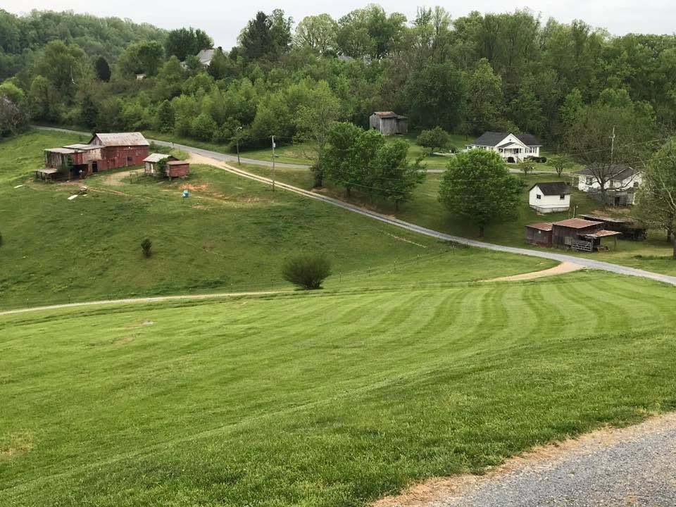 Rolling green hillside with farm buildings, winding dirt road, and trees under an overcast sky