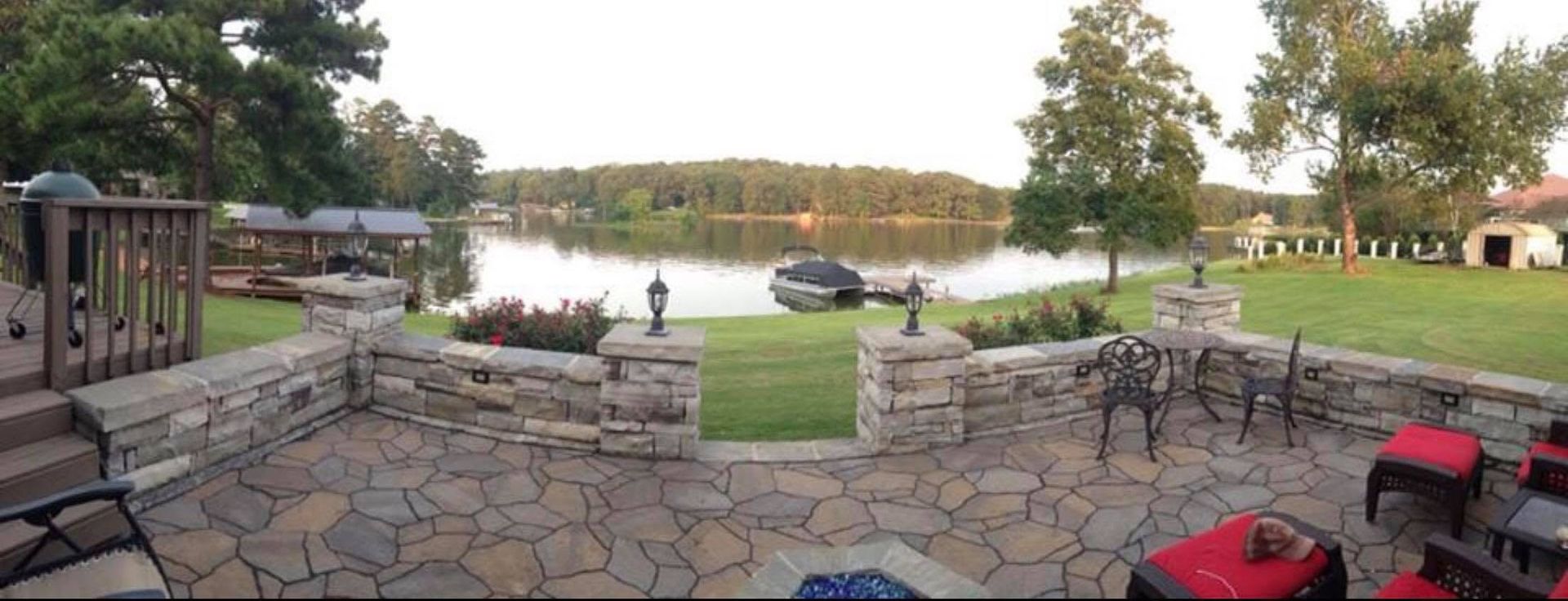 Stone patio overlooking a calm lake with tables, chairs, and a grassy shoreline at sunset