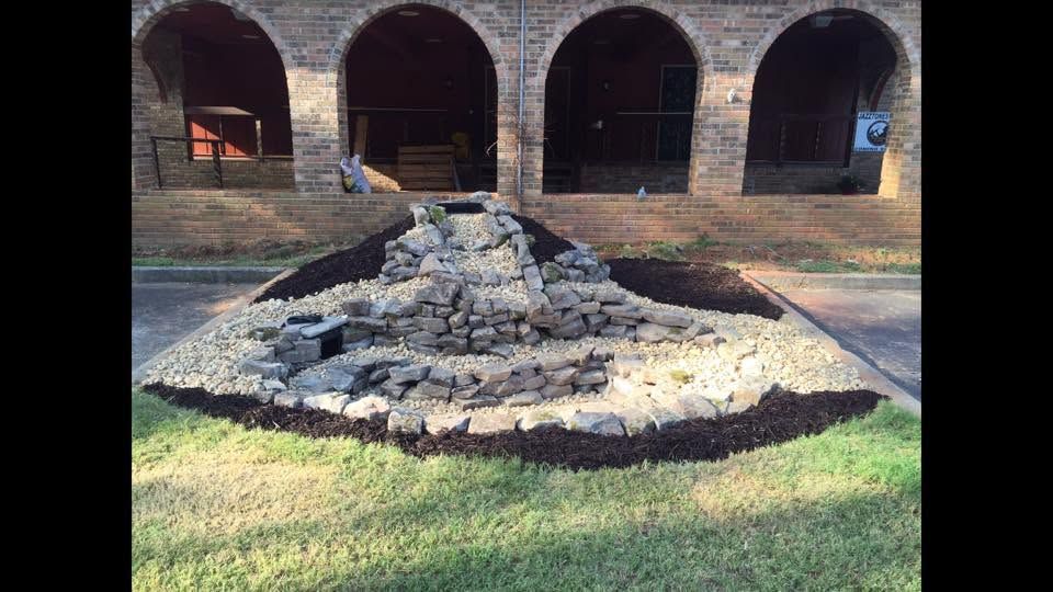 Stone cairn in a grassy courtyard before a brick wall with three arched openings.