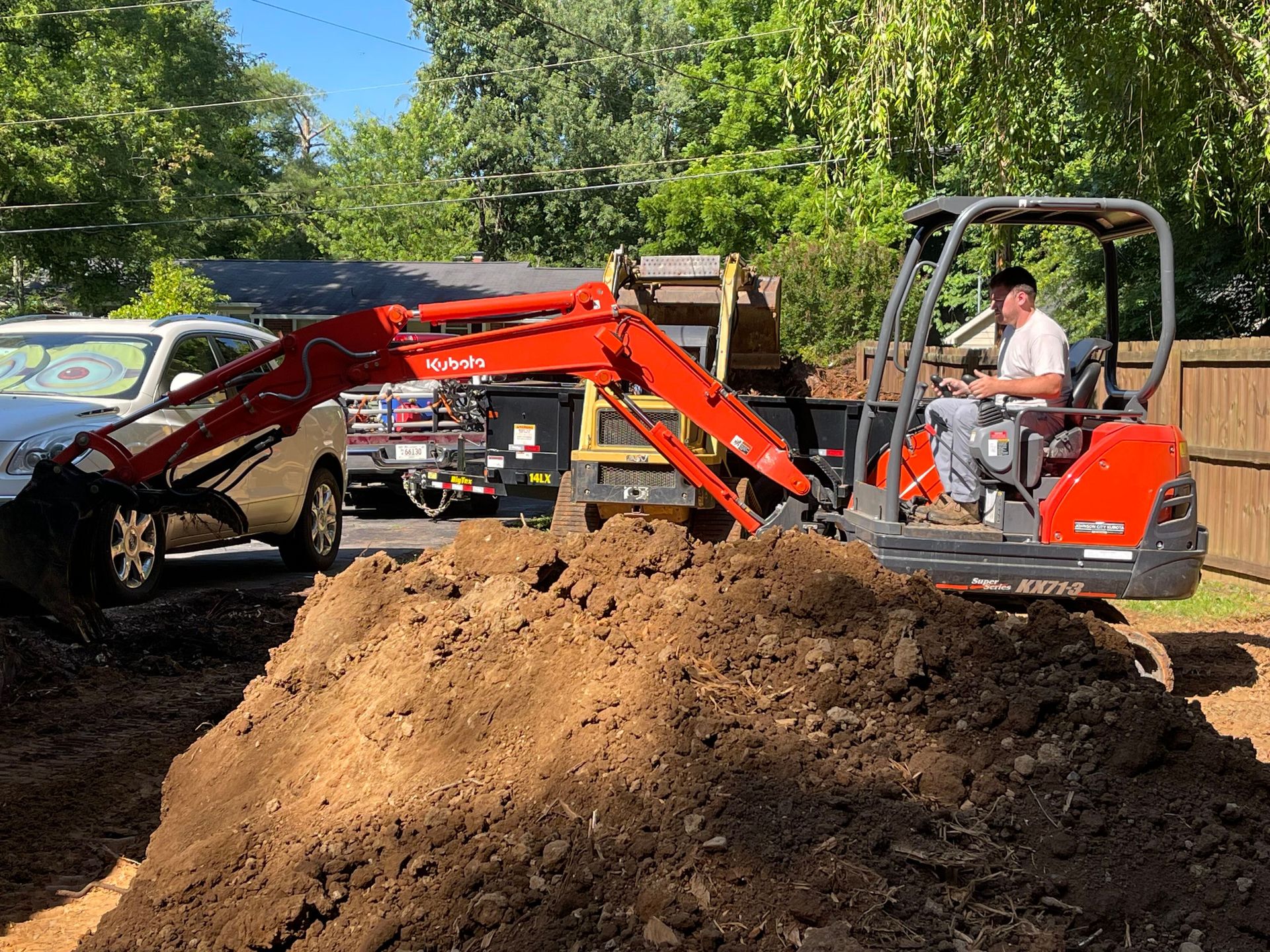 Orange mini excavator moving dirt pile beside parked cars in a wooded area
