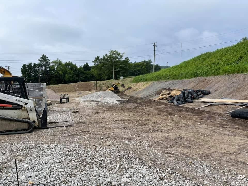 Construction site with gravel, dirt piles, excavator, and overcast sky along a grassy embankment