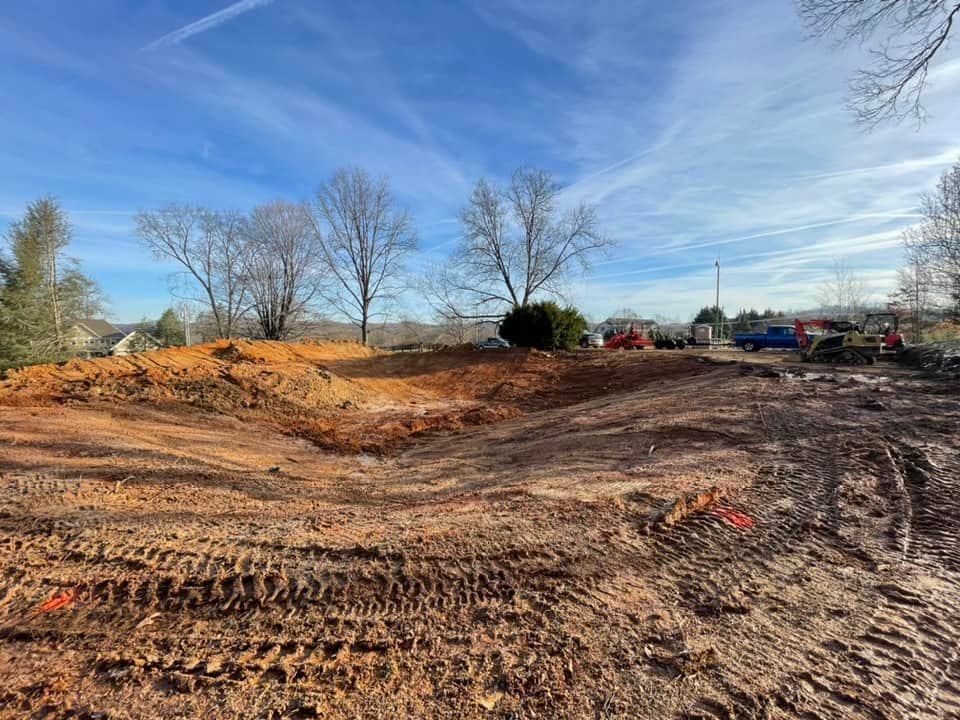 Cleared dirt lot with tire tracks under a blue sky, trees and construction equipment in the distance